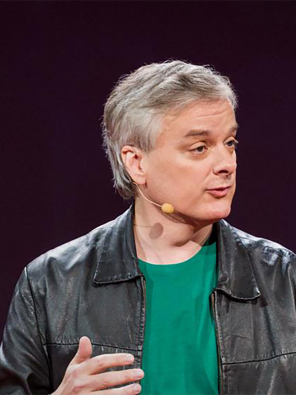 A headshot of David Chalmers, who wears a green shirt and leather jacket, and has a microphone headset.