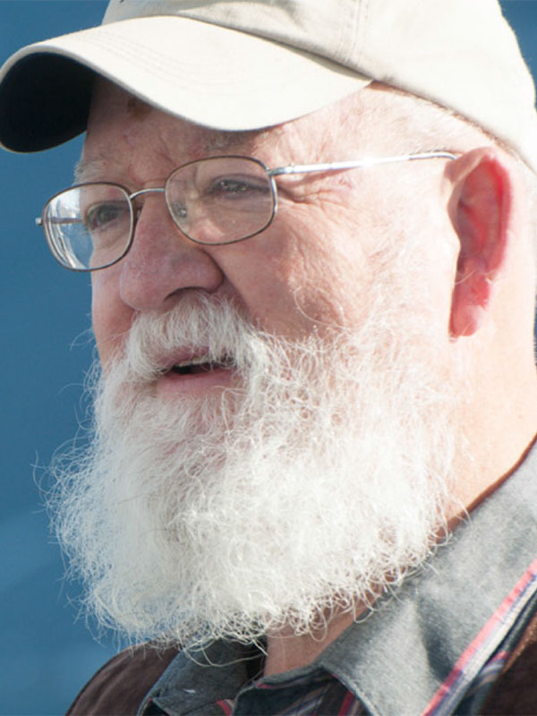 A headshot of Daniel Dennett, who wears a jacket and dress shirt and baseball hat, and poses outside.