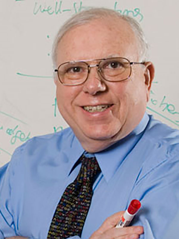 A headshot of J. Michael Dunn, who wears a tie and blue dress shirt and poses in front of a white board.