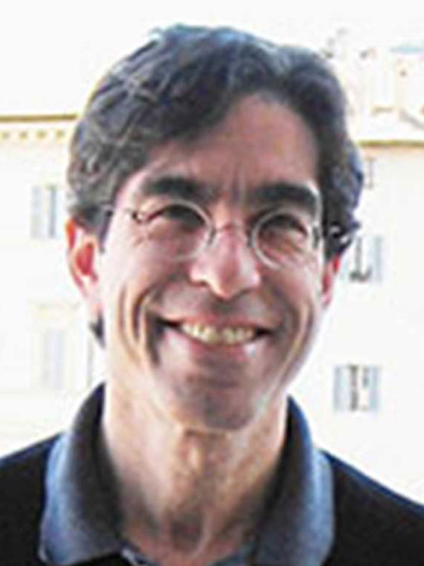 A headshot of Mark Kaplan, who wears a dark blue collared shirt and poses on a city street.