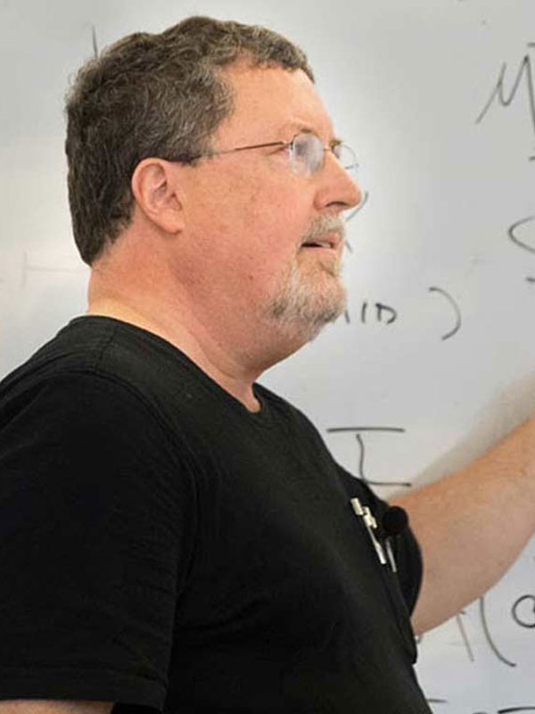 A photo of David McCarty, who wears a black t-shirt and lectures in front of a white board.