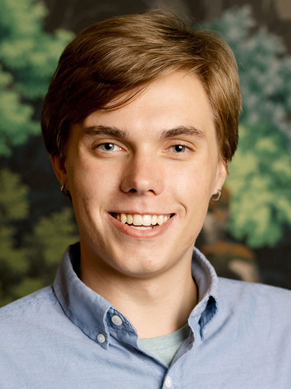 A headshot of Joshua Paschal, who wears a blue dress shirt and poses in front of a green background.