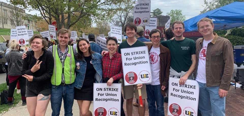 A photo of graduate students on strike on campus, holding various signs in protest that say, "On strike for union recognition."