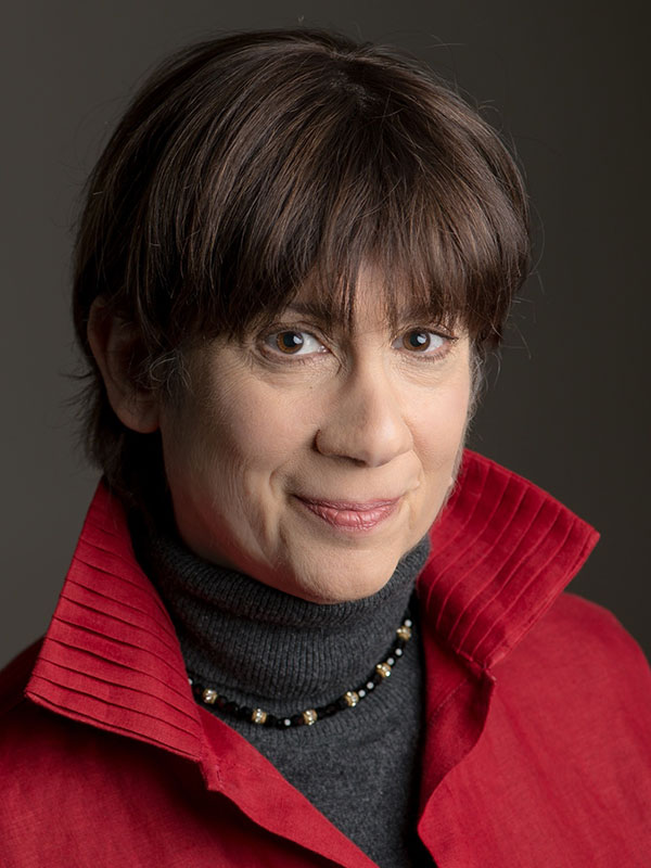 A headshot of Joan Weiner, who wears a red blazer, gray sweater, and poses against a dark background.