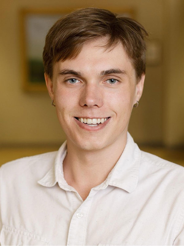 A headshot of Joshua Paschal, who wears a white button-up shirt and poses against a beige background.